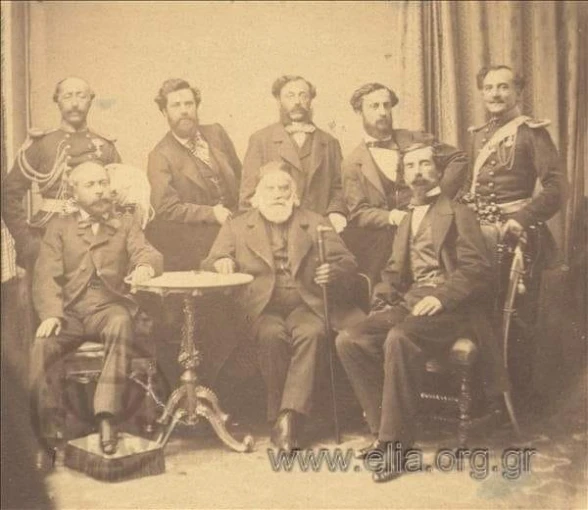 Καθιστοί τα μέλη της επιτροπής: Θρασύβουλος Ζαϊμης, Κωνσταντίνος Κανάρης, Δημήτριος Γρίβας. Όρθιοι τα μέλη της ακολουθίας: Ι. Μόλλας, Δ. Βρατσάνος, Π. Σταύρος, Τ. Φιλήμων, Σπ. Σκουζές.